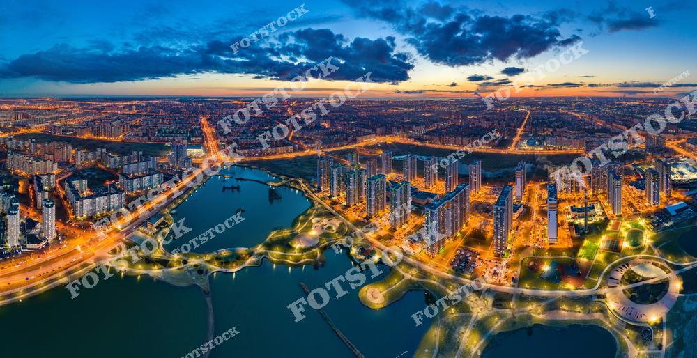 На фотографии изображены современные жилые комплексы Санкт-Петербурга. В центре кадра — высотные дома, окружённые парками, прудами и благоустроенными набережными. Слева проходит крупная магистраль, соединяющая районы. На фоне — бескрайняя городская застройка, уходящая за горизонт. Снимок выполнен в сумерках с воздуха, подчёркивая размах городской инфраструктуры и гармонию с природой.
